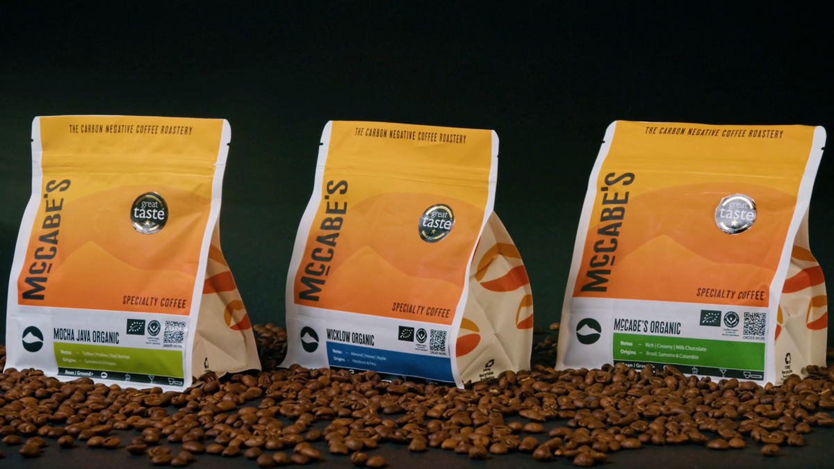 Three bags of McCabe's Specialty Coffee arranged on coffee beans against a dark background. The bags are orange and white with green and blue label accents, and the center bag is partially obscured by a large semi-transparent square overlay.

Details